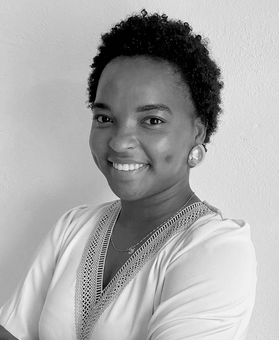 Retrato de Niela Rodrigues com blusa branca com detalhes nas bordas, a sorrir para a câmara diante de uma parede branca