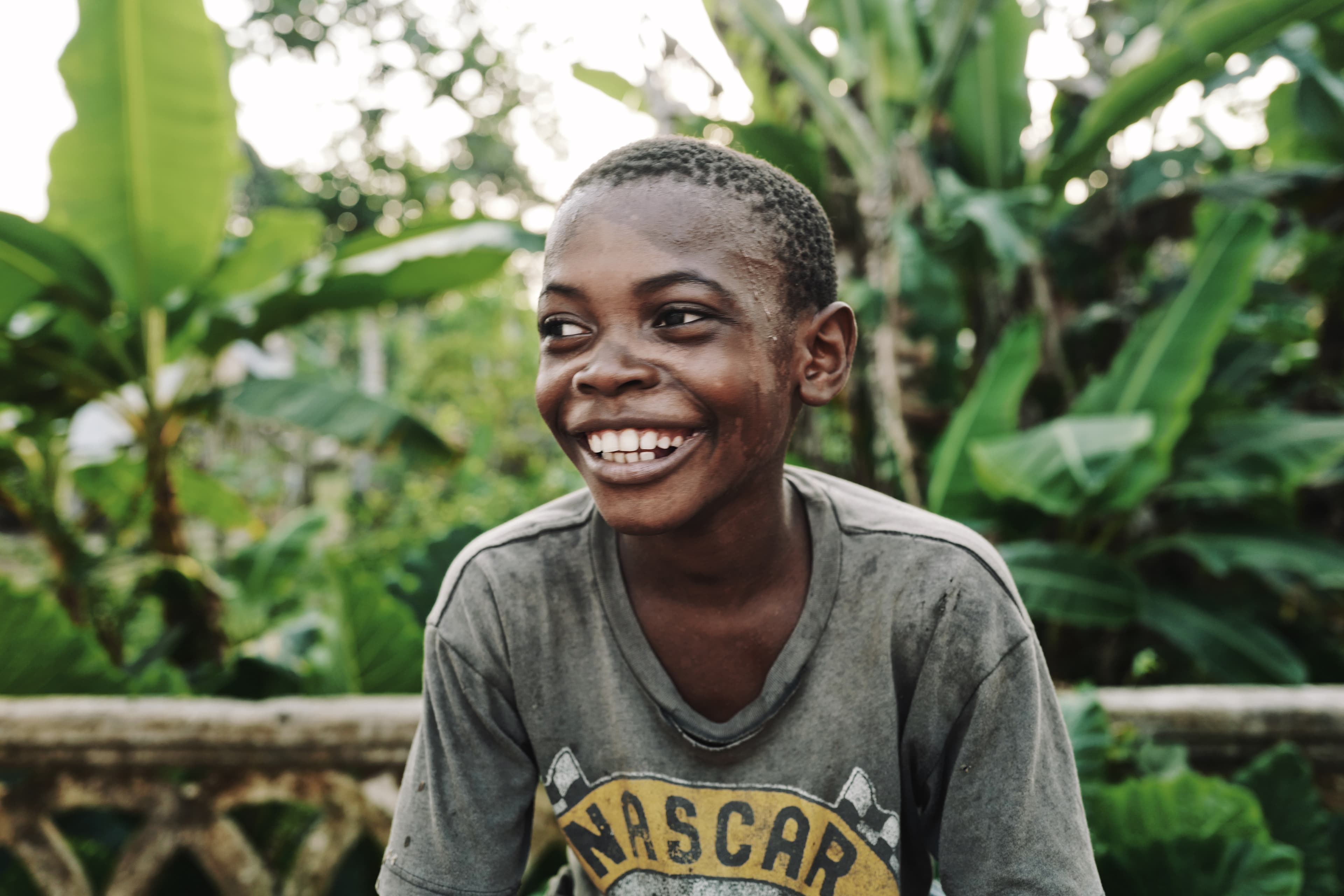 Boy in a grey Nascar T-shirt sits in front of dense green vegetation, smiling broadly