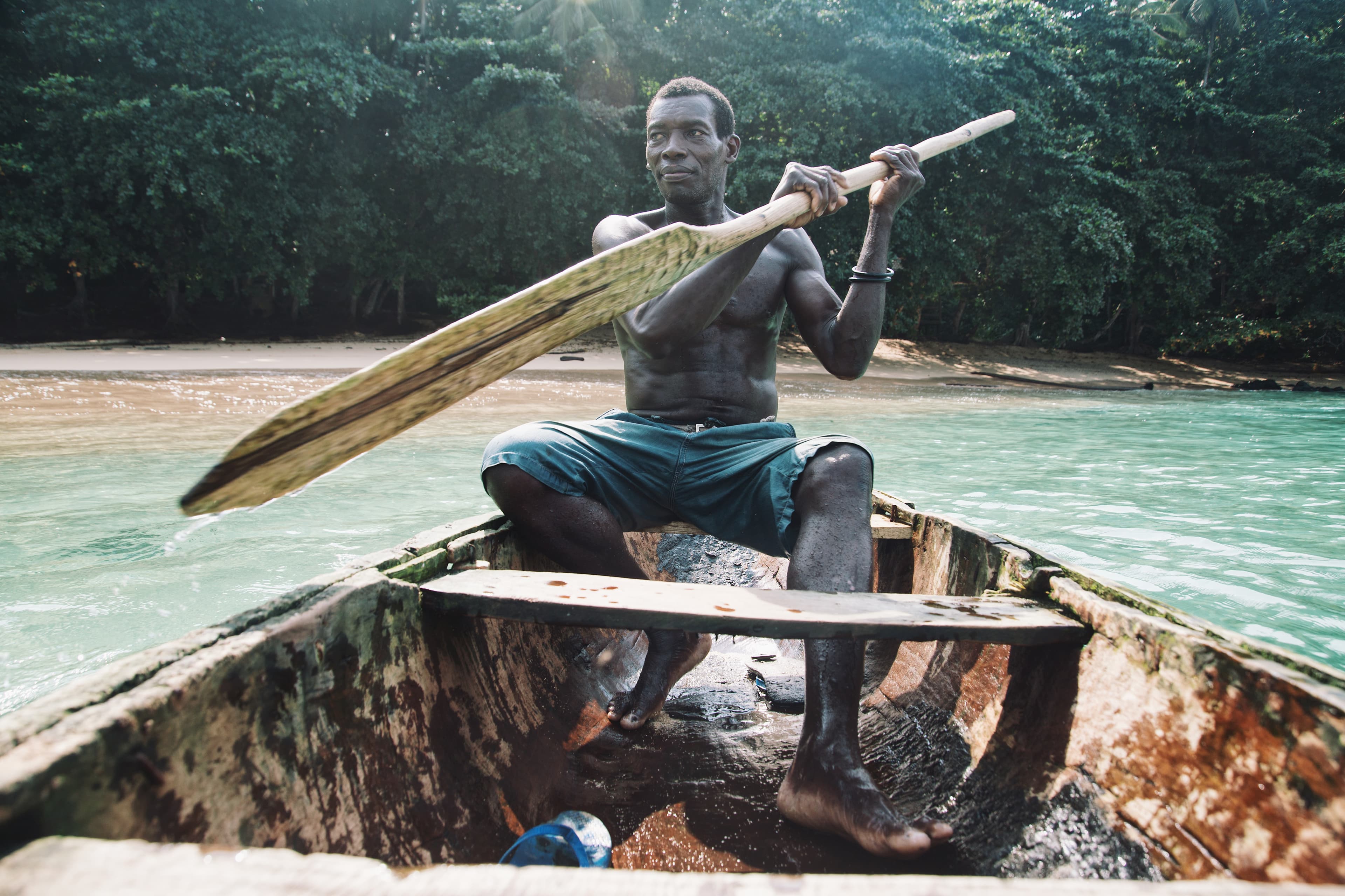 Man sits in a traditional wooden boat with dark blue swim trunks, rowing over turquoise water with a wooden paddle, island in the background